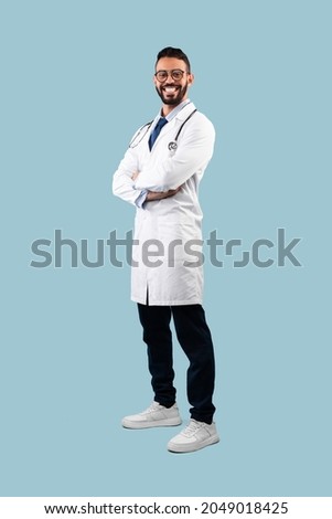 Middle-Eastern Male Doctor Posing Wearing White Uniform Crossing Hands Over Blue Background, Smiling To Camera. Full Length Shot Of Professional Medical Worker, Successful Physician Royalty-Free Stock Photo #2049018425