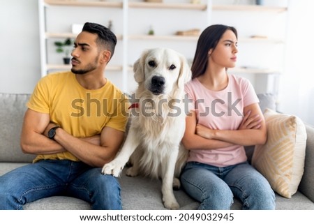 Upset young multinational couple having fight, looking in opposite directions, cute dog sitting on couch between them, indoors. Relationship crisis, marriage conflict, family problems concept Royalty-Free Stock Photo #2049032945