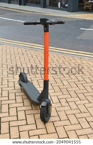 close up of orange and black e-scooter parked on brick pavement in a city alongside tarmac road with double yellow lines.  Electric scooter stood outside with no people