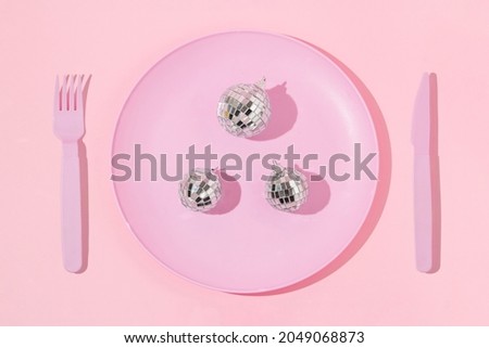Creative layout with pastel pink plate, fork, knife  and disco ball Christmas baubles on pastel pink background. 80s or 90s aesthetic fashion food restaurant concept. Minimal New Year party idea.