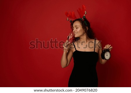 Charming Hispanic woman wearing a deer antler hoop and black velvet dress holding an alarm clock and drinking sparkling wine from a flute with champagne, isolated on red background. Christmas concept