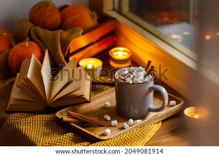 Mug of cocoa or hot chocolate with marshmallows standing next to the window with candles, pumpkins, book and warm blanket. Cozy home atmosphere in rainy autumn day. Нygge lifestyle concept. Royalty-Free Stock Photo #2049081914