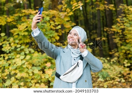 Fashionable young muslim asian girl in hijab taking a selfie on smartphone outdoors in autumn park. Copyspace.