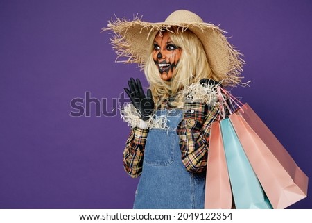 Side view amazed young woman with Halloween makeup mask in straw hat scarecrow costume hold package bags, purchases after shopping isolated on plain dark purple background studio Celebration concept