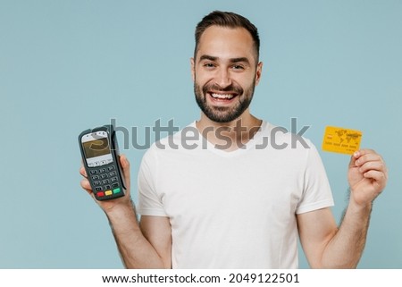 Young happy fun man in blank print design white t-shirt hold wireless modern bank payment terminal to process acquire credit card payments isolated on plain pastel light blue color background studio.