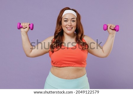 Young fun chubby overweight plus size big fat fit woman 20s wear red top warm up training with female dumbbells raised up hands isolated on purple background home gym Workout sport motivation concept