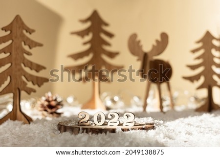 New year 2022. Numbers 2022 on wooden stand on beige pastel blurred background with decorative deer, fir trees, snow and lights. Christmas greeting card.