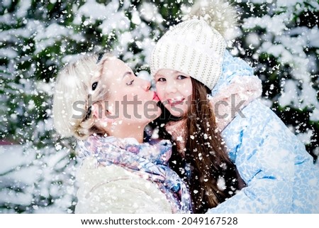 Portrait of a beautiful blonde kissing a little girl on the cheek. Mom and daughter in the winter in the park. Winter forest. Snowfall. Falling snowflakes. A family