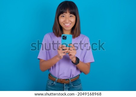 Young beautiful asian girl wearing purple t-shirt over blue background enjoys distant communication, uses mobile phone, surfs fast unlimited internet, has pleasant smile, makes shopping online,