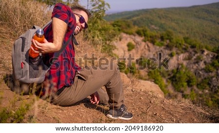 Young man sitting on the cliff and reaching for water at his backpack
