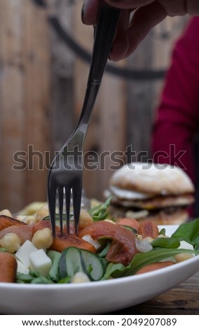 Couple having dinner at the restaurant. Closeup view of a woman holding a fork, eating a fresh colorful salad, and a man having a hamburger with french fries in the background. 