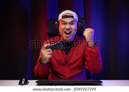 Excited and shocked face of Asian gamer with headphone holding joystick playing video game online sitting on chair at living room. Indian professional gamer streaming on social playing game very fun