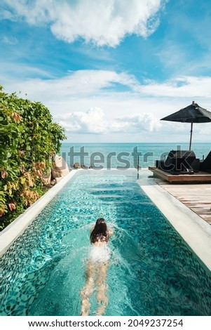 Happy woman in white swimsuit swimming in infinity pool at luxury hotel against ocean front. young female enjoy in tropical resort. Relaxing, summer,  travel, holiday, vacation and weekend concept