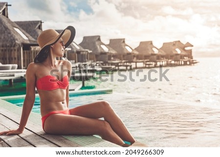 Suntan bikini body Asian woman relaxing sunbathing at luxury vacation in paradise Bora Bora high end resort hotel by the swimming pool at overwater villa suite.