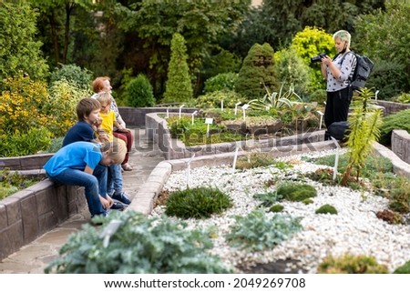 beautiful female photographer with green hair with backpack and camera photographs elderly woman against background of flowers in botanical garden with her beloved grandchildren