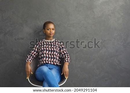 Portrait of young confident African American woman sitting on chair on gray background. Beautiful woman in casual clothes sitting on chair near free space for text looking at camera. Banner.