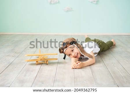 A little boy in aviation helmet lies on the floor next to a wooden toy plane and smiles. The boy dreams of becoming a pilot. Royalty-Free Stock Photo #2049362795