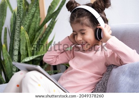 Smiling happy little kid girl using tablet in wireless headphones in couch at home.