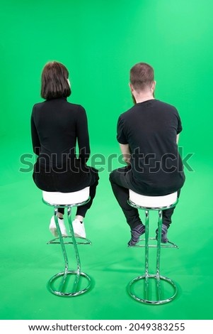 A woman and a man sit with their backs to us on two bar stools in a green screen film studio.