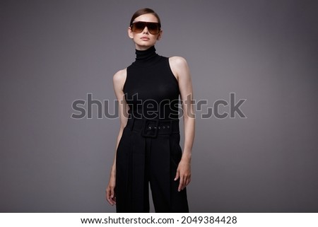 High fashion photo of a beautiful elegant young woman in a pretty black pants, sleeveless top, stylish sunglasses posing over gray background. Slim figure. Studio Shot. 