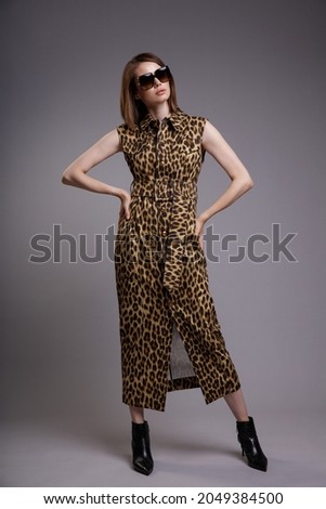 High fashion photo of a beautiful elegant young woman in a pretty shirt dress sleeveless animal leopard print, black ankle boots, stylish sunglasses posing over gray background. Slim figure.