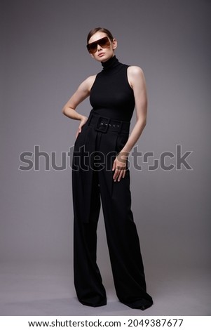 High fashion photo of a beautiful elegant young woman in a pretty black pants, sleeveless top, stylish sunglasses posing over gray background. Slim figure. Studio Shot. 