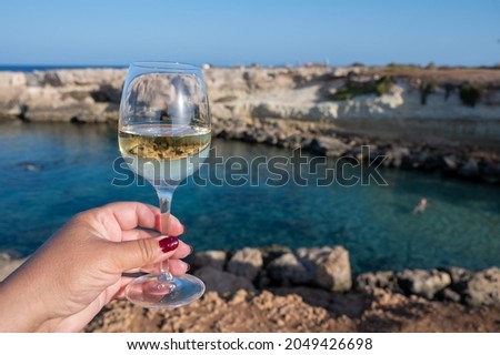 Woman's hand holding glass of white dry white wine with view on rocks and blue sea bay water near Protaras touristic town on Cyprus, party time