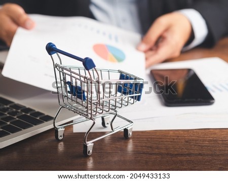 A mini shopping trolley on a wooden table with a businessman holding a document while sitting in the office with blur background. Business and e-commerce concept.