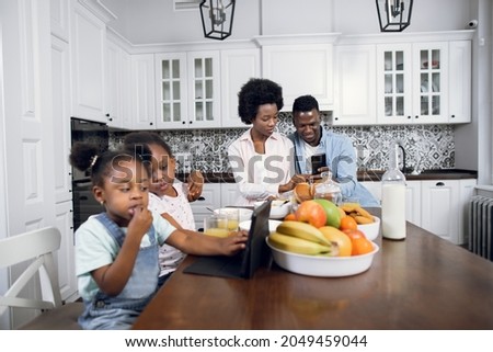 Two african sisters sitting on chairs and watching cartoons on digital tablet while their young parents using smartphone on background. Family with gadgets on kitchen. Modern lifestyles.