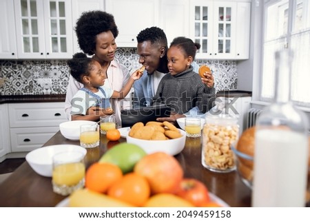 Beautiful african american family of four using digital tablet during breakfast on bright kitchen. Happy young parents watching cartoons with their two daughters while eating at home.