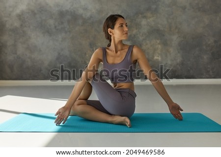 Spine twisting pose. Woman sitting on blue mat in Ardha Matsyendrasana yoga position with straight arms down and opened palms, head turned to left, legs to right, having relaxed face expression