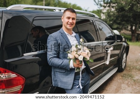 A handsome, young, smiling groom stands near a black decorated car with a bouquet of flowers in his hands. Wedding photography, portrait.