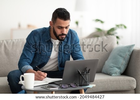 Serious young Arab man using laptop, working remotely, taking notes during remote business meeting from home. Millennial Eastern guy studying online, writing down info. Modern technologies concept