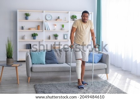 Young black guy with plastered leg walking with crutches, suffering from pain at home, copy space. Full length portrait of millennial Afro man having fractured limb. Health care concept Royalty-Free Stock Photo #2049484610
