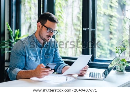 Serious pensive thoughtful focused young casual entrepreneur small business owner accountant bookkeeper in office looking at and working with laptop and income tax return papers and documents Royalty-Free Stock Photo #2049487691