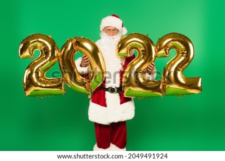Santa claus in costume holding balloons in shape of 2022 isolated on green