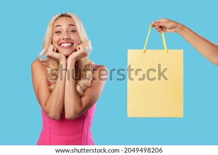 Gift Delivery Concept. Excited millennial woman waiting for surprise, hand holding and giving paper package bag with blank empty space for mock up design, isolated on blue studio background Royalty-Free Stock Photo #2049498206