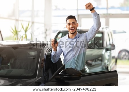 Emotional handsome middle-eastern guy raising hand up and smiling at camera, showing key from his brand new black car, copy space. Rich arabic man buying auto at luxury dealership salon Royalty-Free Stock Photo #2049498221