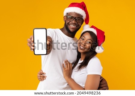Great Application. African American Couple Showing Smartphone Empty Screen Wearing Santa Hats Advertising Christmas Offer Or Message Standing Over Yellow Background. Selective Focus On Phone