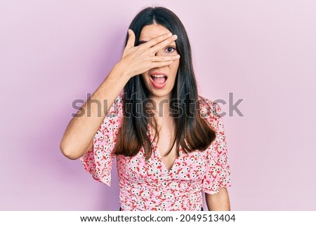 Young brunette woman wearing summer dress peeking in shock covering face and eyes with hand, looking through fingers with embarrassed expression. 