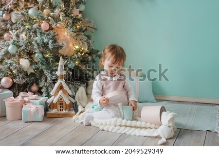 Little girl opens gifts while sitting under the Christmas tree. Christmas interior in pink and blue colors.