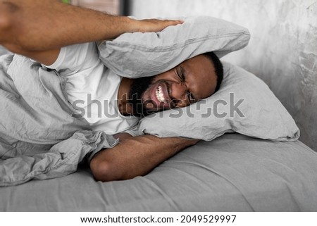 I Can't Sleep. Portrait of irritated young black man lying in bed and covering ears with pillows, hearing and suffering from too loud sound. Angry millennial guy tired of noisy neighbors Royalty-Free Stock Photo #2049529997