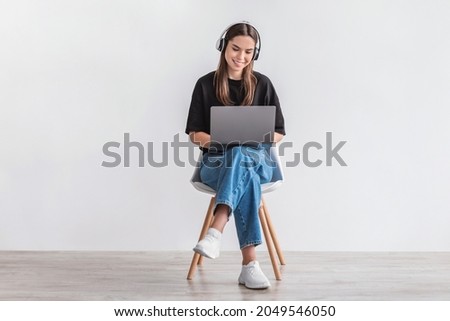 Cheery Caucasian woman in headphones having online video call on laptop, sitting on chair against white wall, full length. Millennial lady participating in webinar, communicating remotely on internet
