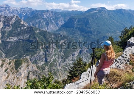A young woman traveler stands on top of a mountain looking down at the Grlo Sokolovo Gorge and the surrounding mountains, next to an action camera on a tripod, capturing a breathtaking time-lapse view