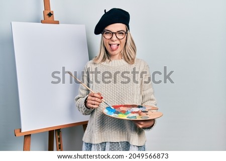 Beautiful caucasian woman drawing with palette on easel stand sticking tongue out happy with funny expression. 