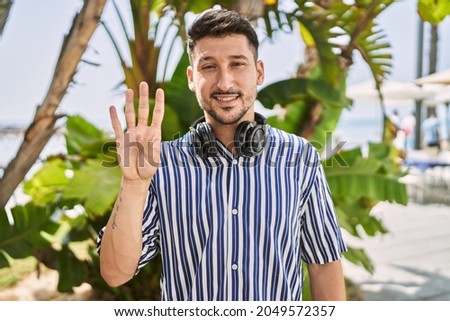 Young handsome man listening to music using headphones outdoors showing and pointing up with fingers number four while smiling confident and happy. 