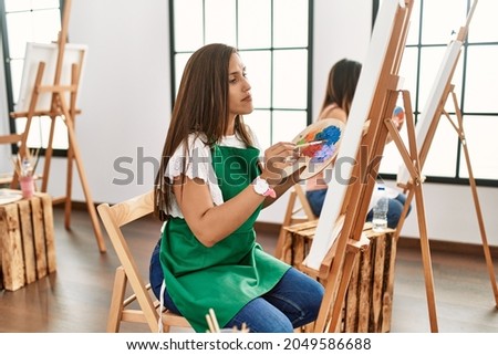 Young hispanic artist women painting on canvas at art studio relaxed with serious expression on face. simple and natural looking at the camera. 