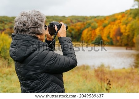 Seniors and technology, retirement hobby concept. Back view senior woman takes photo on digital camera, elderly woman tourist takes pictures of autumn beautiful park and lake, copy space.