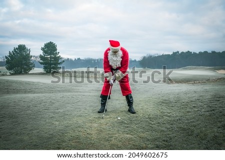 Santa Claus in a red traditional costume plays golf on a green frosty field, outside in winter. Royalty-Free Stock Photo #2049602675