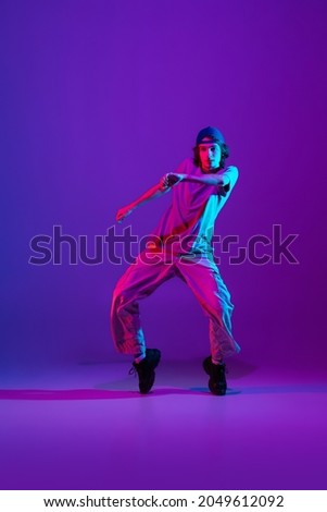 In action, motion. Young stylish man, break dancing, hip-hop dancer practicing solo in casual clothes isolated over purple background in neon pink light at dance hall. Youth culture, movement, style